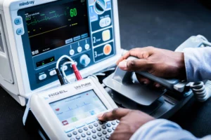 Manos de un técnico médico manejando un desfibrilador y monitor de signos vitales, representando el monitoreo y la tecnovigilancia en equipos biomédicos.